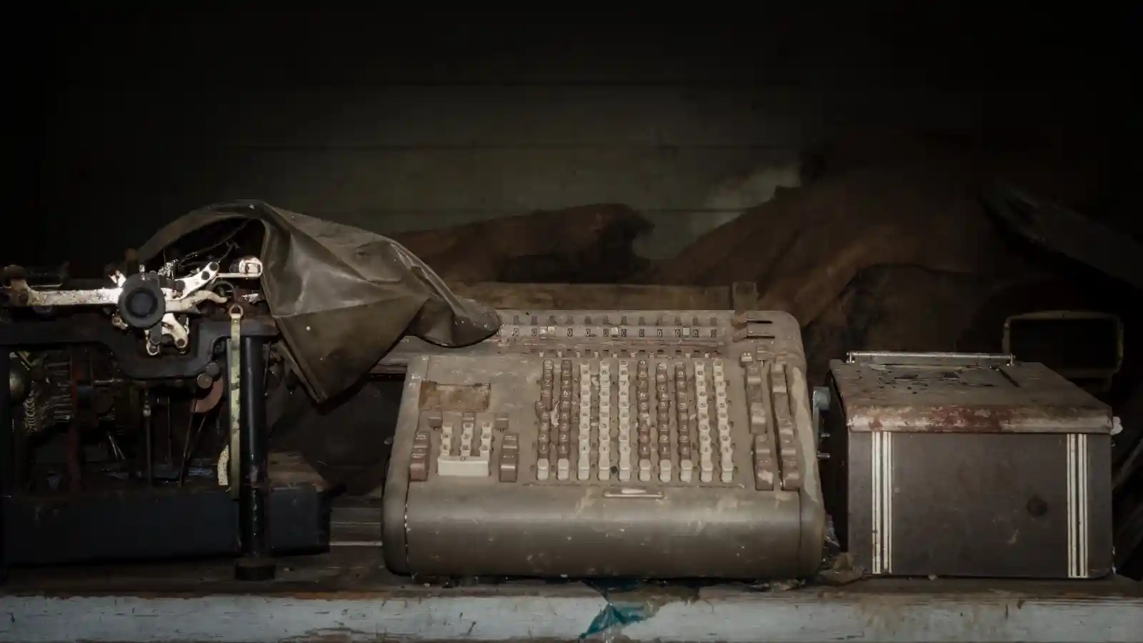 An abandoned adding machine sits in shadowed silence, covered in a thick coat of dust, keys untouched for years. To its left, a skeletal typewriter; to its right, a rusted, closed box. The room is heavy with the weight of forgotten tales, each machine a relic of untold stories, of numbers added up but never spoken aloud. The past is held captive here, where machines once whispered secrets in the dark.